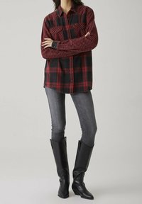 Femme portant une chemise à carreaux rouge et noir, un jean skinny gris et des bottes en cuir noir jusqu'aux genoux, se tenant avec les bras croisés.