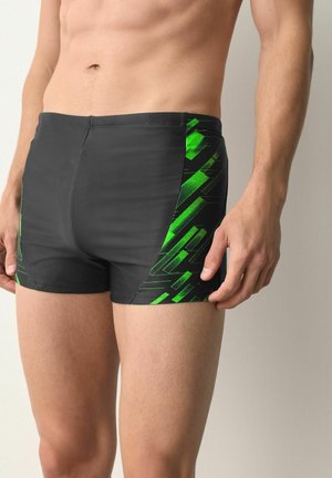 Male torso wearing black swim trunks with green geometric side panels, standing against a neutral light background.
