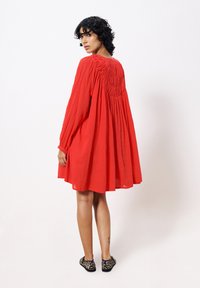 Rotes knie-langes Kleid mit langen Ärmeln, strukturiertem Oberteil und gerafftem Rückendetail. Kombiniert mit Hausschuhen im Tierdruck.