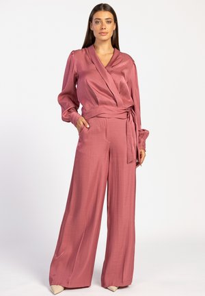 Roze satijnen wikkeltop en set met wijde pijpen. Heeft lange mouwen en een strik in de taille. Soepele textuur, losse pasvorm en minimale accenten.