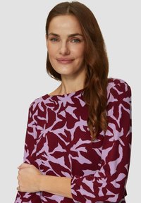 Bordeauxrotes Kleid mit lila abstraktem Muster; runder Ausschnitt, Dreiviertelärmel und weicher Stoff. Das Model steht mit verschränkten Armen vor einem neutralen Hintergrund.