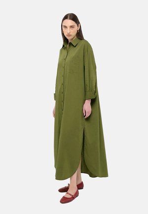 Femme aux longs cheveux bruns porte une longue robe boutonnée vert olive avec des fentes latérales et des chaussures plates rouges, debout devant un fond uni.