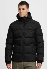 Black puffer jacket with a high collar and attached hood. Features quilted design, side pockets, and a circular logo on the chest.