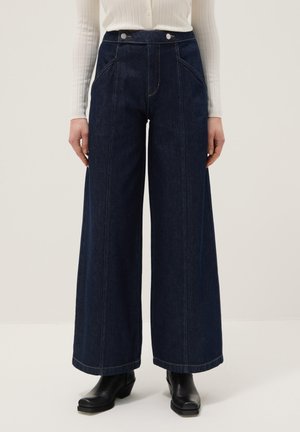 Jeans de mezclilla azul oscuro de pierna ancha con costuras frontales, cintura alta, bolsillos laterales, botones plateados, combinados con botas negras y un suéter blanco de canalé.