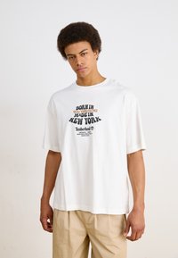 Hvid bomulds t-shirt med korte ærmer, prydet med fed trykt tekst i sort og orange. Rund hals design og afslappet pasform. Timberland logo inkluderet.