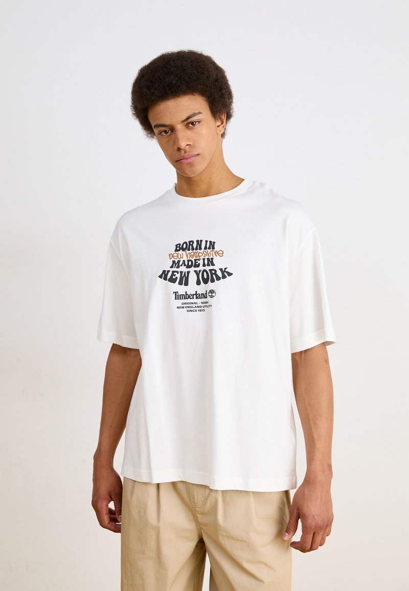 Hvid bomulds t-shirt med korte ærmer, prydet med fed trykt tekst i sort og orange. Rund hals design og afslappet pasform. Timberland logo inkluderet.
