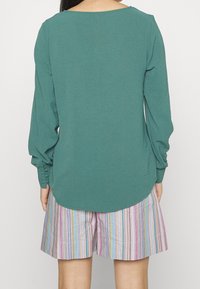 Žena ve svetlemodrém dlouhém rukávu s manžetami na knoflíky a barevnými pruhovanými krátkými kalhotami, stojící čelem k bílé pozadí.