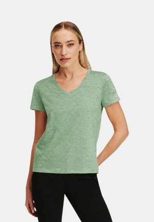 Donna con capelli biondi lisci che indossa una maglietta verde chiaro a maniche corte con scollo a V e pantaloni neri, in piedi con una mano sul fianco.