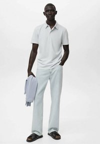 Ljusgrå kortärmad pikétröja, ljusblå jeans och svarta sandaler. Håller en ljusblå tröja och står mot en enfärgad bakgrund.