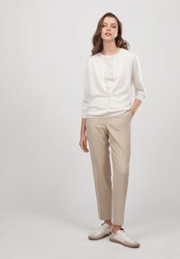 Jasno beżowy kardigan na białej bluzce, zestawiony z beżowymi spodniami i białymi sneakersami. Gładka tekstura, dopasowany krój oraz subtelny detal w postaci guzików.