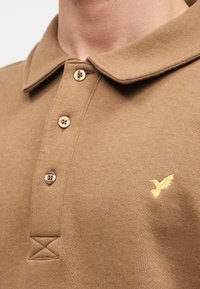 Καφέ πόλο μπλούζα από βαμβάκι, με κλασικό γιακά, τρία κουμπιά και κεντητό κίτρινο λογότυπο πουλιού στο στήθος.