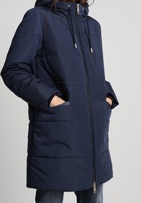 Abrigo acolchado azul marino con capucha, que cuenta con un cierre frontal, bolsillos laterales y una textura acolchada. Terminado con cordones ajustables.