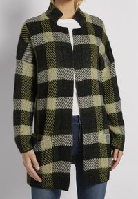 Zeleno-černý kostkovaný kardigan s vysokým límcem, otevřeným předním dílem a kapsami. Vyroben z texturované tkaniny, s kostkovanými vzory v různých velikostech.