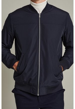 Homme portant une veste bomber bleu marine avec fermeture éclair devant et les mains dans les poches, associée à un col roulé blanc et un pantalon bleu marine.