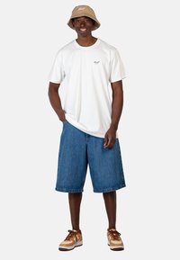 Camiseta de algodón blanca con un pequeño logo, pantalones cortos de mezclilla azules y un sombrero tipo bucket beige. Zapatillas marrones con suelas blancas completan el atuendo.