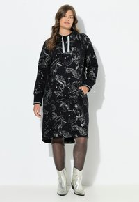 Schwarzes Kapuzenkleid mit silbernen, meerinspirierten Mustern, langen �Ärmeln, zwei Taschen und kontrastierenden grauen Kordeln. Getragen mit silbernen Stiefeletten.