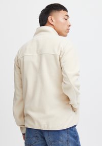 Beige fleecejakke med høy krage, strukturert materiale og elastisk kant. Har en ensfarget bakside med en subtil søm over skuldrene.