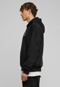 Mustapaita, jossa on huppu, etutasku, löysä istuvuus ja kirjailtu graafinen kuvio. Varren päällä on ribbireunat ja rento muotoilu. Kangas näyttää pehmeältä.