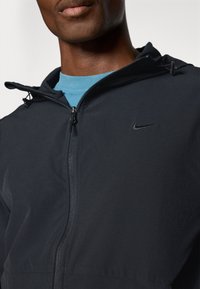 Homme portant une veste noire à fermeture éclair avec une capuche, sur un t-shirt bleu, montrant la partie supérieure de la poitrine et du cou.