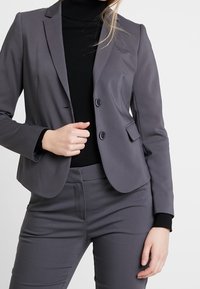 Blazer gris sur mesure avec deux boutons, col à revers et poches plaquées, associé à un pantalon gris assorti et un col roulé noir.