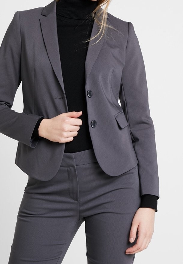 Blazer gris sur mesure avec deux boutons, col à revers et poches plaquées, associé à un pantalon gris assorti et un col roulé noir.