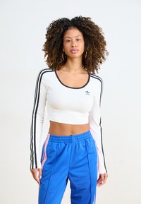 White long-sleeve crop top with black collar and three black stripes on sleeves. Paired with blue pants featuring pink side stripes.