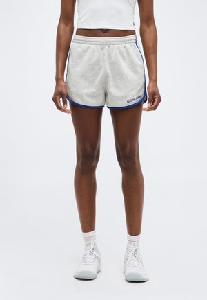 Person iført lysegrå sportsshorts med blå kant og hvide sneakers, stående mod en ensartet lys baggrund.