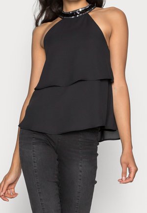 Vrouw draagt een mouwloze zwarte gelaagde top met een halternek met pailletten, gecombineerd met donkergrijze strakke jeans.