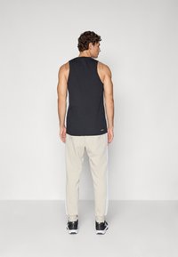 Vyras stovi nugarą, apsirengęs juodu be Rankovių marškinėliais, smėlio spalvos treningų kelnėmis su baltomis juostelėmis ir juodais sportiniais batais ant vientiso fono.