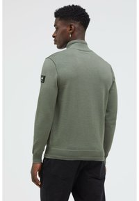 Olivgrüner Strickpullover mit hohem Kragen, gerippten Bündchen und Saum. Verfügt über ein schwarzes Logo-Patch am linken Ärmel.