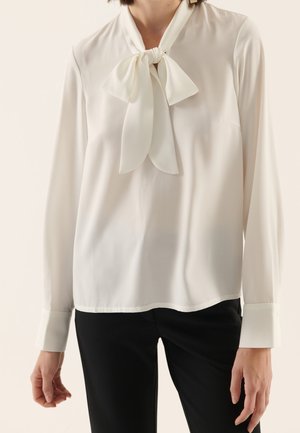 Femme portant un chemisier blanc à manches longues avec un nœud noué au col et un pantalon noir sur un fond uni.
