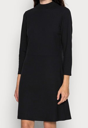 Woman wearing a long-sleeve black dress with a high ribbed neckline standing against a plain background.
