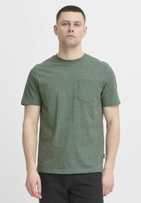 Camiseta de algodón verde con una textura moteada, mangas cortas, un escote redondeado y un bolsillo en el pecho. Diseño minimalista con corte recto.