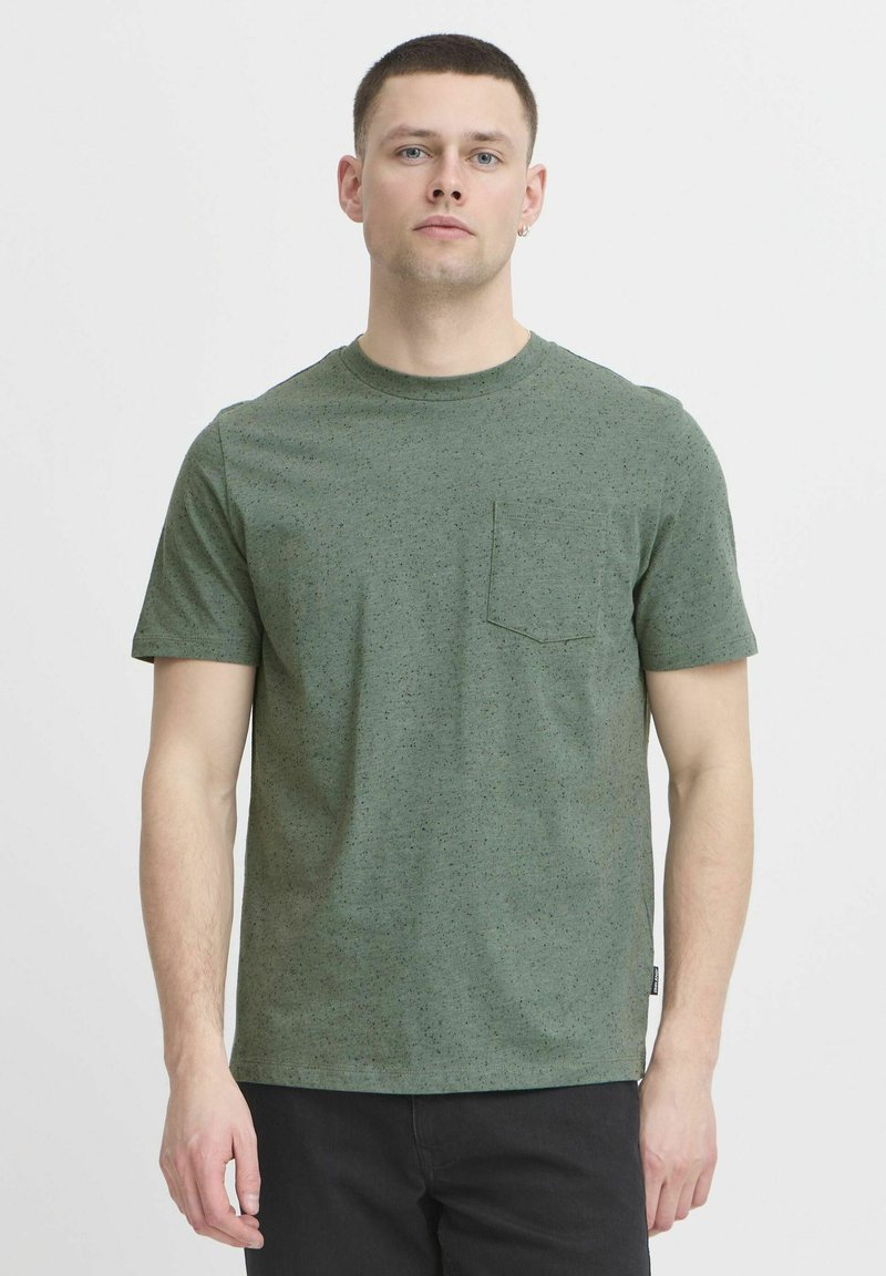 Camiseta de algodón verde con una textura moteada, mangas cortas, un escote redondeado y un bolsillo en el pecho. Diseño minimalista con corte recto.