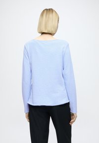 Jasnoniebieski top z długim rękawem z miękkiej tkaniny, o szerokim dekolcie i zakrzywionym dole, zestawiony z ciemnymi spodniami. Prosty, minimalistyczny design.