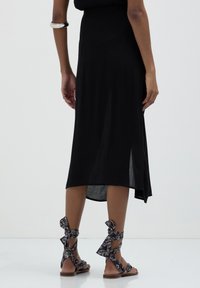 Femme portant une robe midi noire et des sandales plates à motifs noirs, se tenant sur un sol blanc réfléchissant.