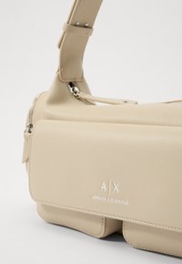 Beżowa torebka na ramię z ekoskóry z zamkiem błyskawicznym, dwoma przegrodami oraz wytłoczonym logo "A/X ARMANI EXCHANGE" z przodu. Regulowany pasek.