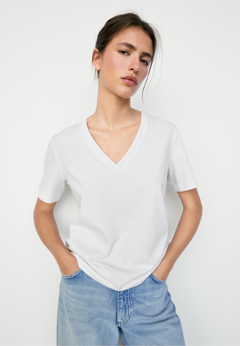 Frau mit braunem, zurückgebundenem Haar, die ein weißes T-Shirt mit V-Ausschnitt und kurzen Ärmeln sowie hellblaue Jeans trägt und vor einem schlichten weißen Hintergrund steht.
