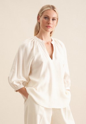 Top bianco, morbido e svasato con scollatura a V, maniche lunghe a sbuffo e dettagli arricciati sulle spalle. Realizzato in un tessuto leggero e soffice.