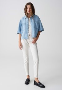 Camicia in denim azzurro chiaro, oversize, con maniche corte, due tasche frontali, t-shirt bianca, pantaloni bianchi slim e mocassini neri. Sfondo neutro.