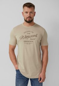Beige T-shirt met korte mouwen, voorzien van een ronde hals en een grafische print in bruine tekst, gecombineerd met blauwe denim jeans.