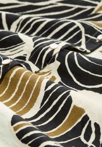 Tessuto di cotone con un audace motivo a foglie in nero, crema e giallo senape, con bottoni bianchi lungo la patta.