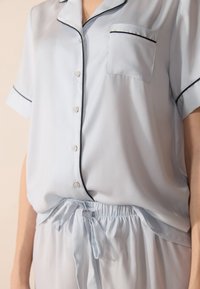 Un ensemble de pyjama en soie bleu clair comprenant un haut à boutons avec des accents de piping noirs et une poche poitrine ; taille élastique avec un détail à nouer.