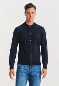 Mężczyzna w granatowym swetrze zapinanym na guziki z ribbed detalami i niebieskich dżinsach, stojący na tle jasnym.