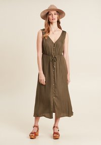 Olivgrünes Kleid mit V-Ausschnitt, Knopfleiste, Bindegürtel und knielangem Saum. Getragen mit einem beigefarbenen Hut und braunen Keilsandalen. Glatte Textur.