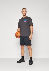Šedé tričko s krátkým rukávem a barevným potiskem, doplněné černými síťovanými basketbalovými šortkami a černými teniskami, držící pomerančový basketbal.