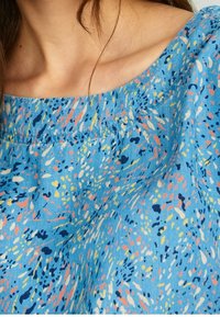 Haut en tissu bleu avec un motif éclaboussé en rose, jaune et noir. L'encolure est large avec des détails froncés près des épaules.