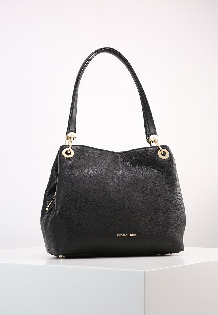 Sac à main en cuir noir texturé Michael Kors avec des ferrures dorées et doubles anses d’épaule sur un socle blanc contre un fond clair.