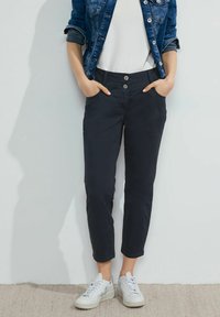 Marina cropped byxor med två framfickor, knappgylf och en slät tygtextur, tillsammans med vita sneakers.