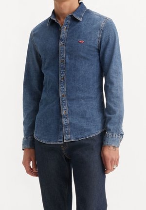 Homme portant une chemise en denim bleue Levi's à manches longues et un jean bleu foncé, debout devant un fond blanc.